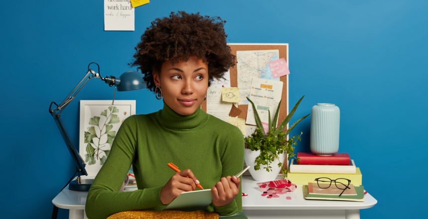 Pensive curly young girl writes down future plans and goals in notepad, thinks about good idea, poses against cozy workplace interior, dressed in comfortable green jumper, isolated on blue wall